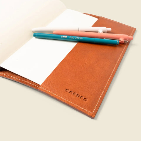 CASUPO’s Refillable A5 Leather Notebook Cover – Tan Limited Edition holds a white notebook and four pens in white, blue, and pink tones—neatly arranged on a light surface.