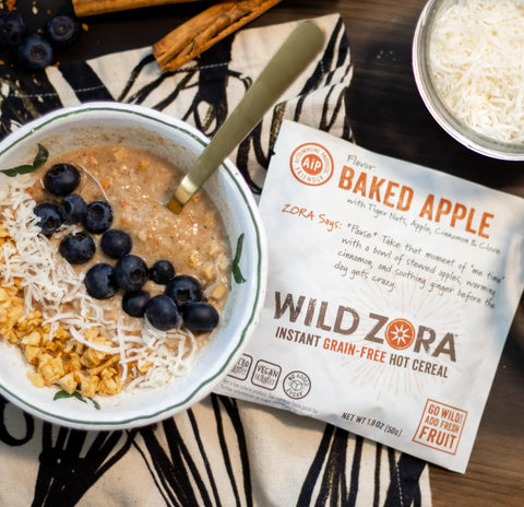 A bowl of Wild Zora Instant Cereal - AIP Baked Apple With Tiger Nuts, Apple, Cinnamon & Clove is topped with shredded coconut, blueberries, and diced apples on a striped cloth beside its BULK 1.5 lb. packet, with coconut and cinnamon sticks nearby.