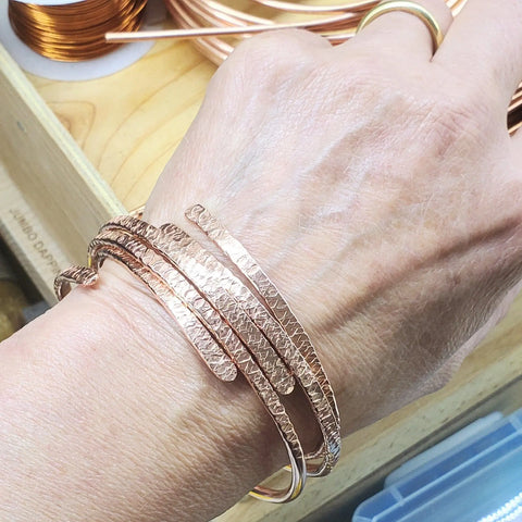 A hand wears the Alexa Martha Designs Adjustable 10 Gauge Copper Hammer Textured Overlap Bangle, a stunning copper anniversary gift, with copper wire coils and crafting tools in the background.