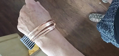 Someone wears an Alexa Martha Designs Adjustable 10 Gauge Copper Hammer Textured Overlap Bangle on their wrist, standing on a wooden floor; part of their patterned skirt and one shoe are visible—a stylish choice for unique copper anniversary gifts.
