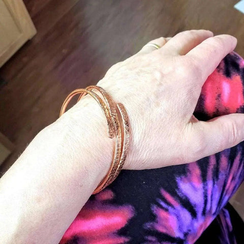A hand wears the Alexa Martha Designs Adjustable Hammered Copper Overlap Bangle, resting on a leg in vibrant pink, purple, and black tie-dye leggings. A wooden floor and a cabinet corner are in the background.