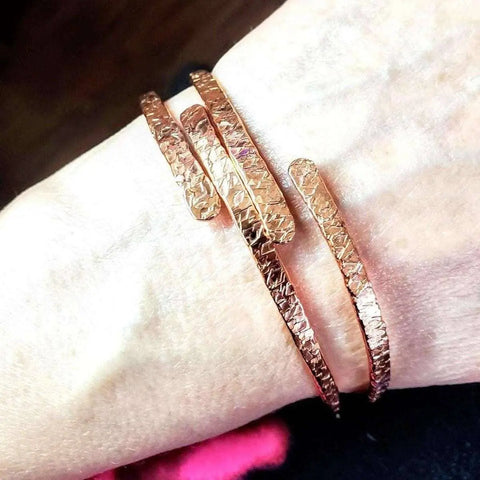 A close-up shows a wrist wearing the Alexa Martha Designs Adjustable Hammered Copper Overlap Bangle for Him or Her, featuring a textured, open-ended design against a blurred brown background.