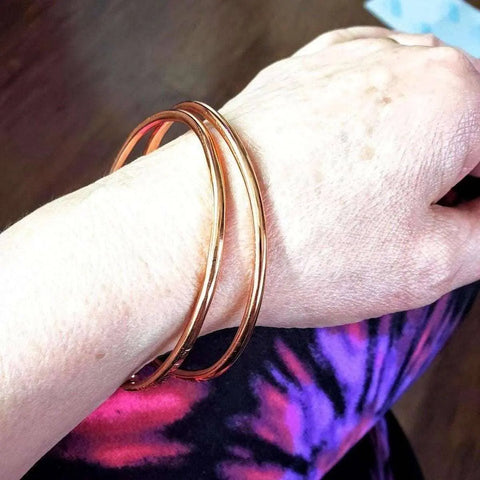 A hand models the Alexa Martha Designs Adjustable Hammered Copper Overlap Bangle, resting on leggings with a pink, purple, and black tie-dye pattern. The background is blurred.