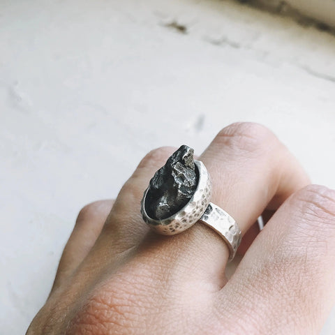 A hand displays Yugen Handmade’s Oval Raw Meteorite Ring in Silver, featuring a rough black stone in a textured hammered band, set against a softly blurred background.