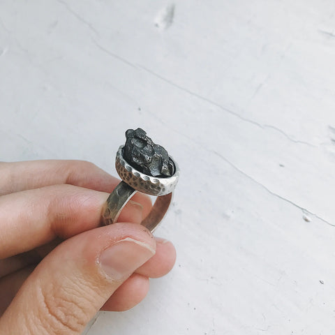 A hand holds the Yugen Handmade Oval Raw Meteorite Ring in Silver, featuring a hammered band and bezel-set rough black meteorite, against a white textured background.