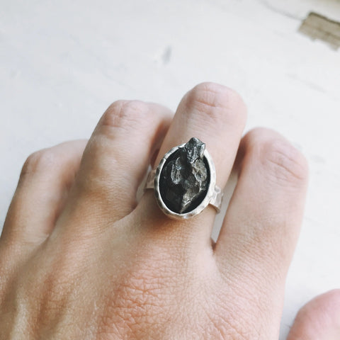 A hand displays Yugen Handmade’s Oval Raw Meteorite Ring in Silver, featuring a striking rough black stone set in an oval bezel against a light backdrop—a perfect piece for outer space jewelry enthusiasts.