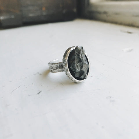 The Yugen Handmade Oval Raw Meteorite Ring in Silver features a rough, dark stone setting accented by soft natural light, highlighting its textured band and unique, rustic outer space-inspired design on a white surface.