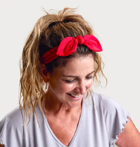 A smiling woman with wavy light brown hair in a ponytail wears the Goodnighties Knotted Bow Headband in bright red, paired with a light gray top, while looking down against a plain light background.