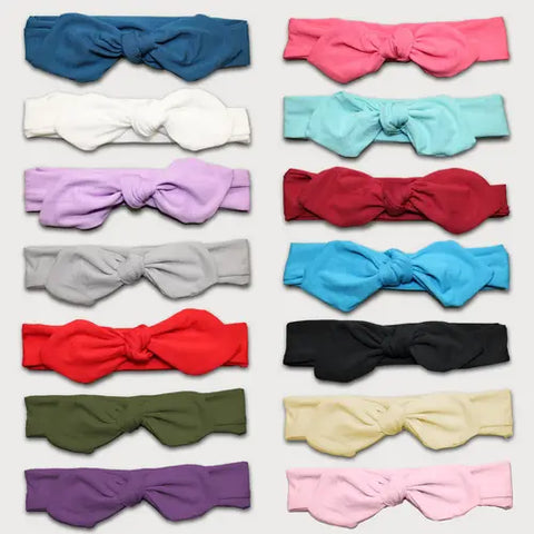 A display of 15 Goodnighties Knotted Bow Headbands, each with a large bow, arranged in 3 columns and 5 rows. These adjustable, solid-color headbands are handmade in the USA in blue, pink, lilac, green, and more.