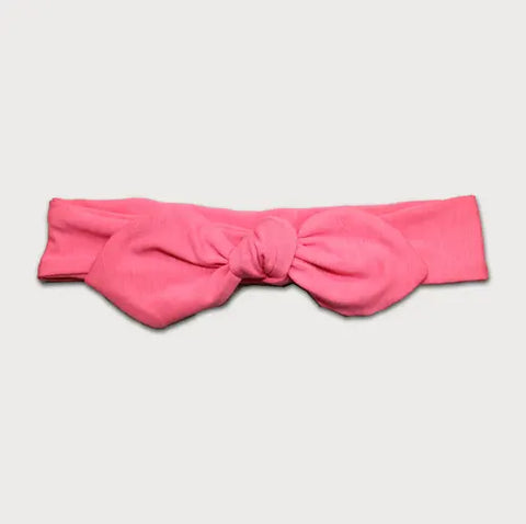 The Goodnighties Knotted Bow Headband in pink features a large center knot and bow, laid flat on a light background. This adjustable headband blends style with comfort.