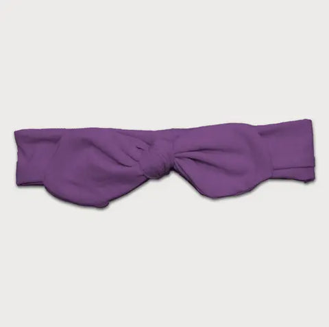 The Goodnighties Knotted Bow Headband features a wide, purple design with a large front knot, shown flat on a white background. Handmade in the USA.