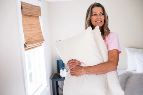 A smiling woman with long hair, wearing a pink shirt, stands in a bright bedroom and hugs the American Blossom Linens Natural Cotton Pillow With Wool Filling. A window with bamboo shade and a bedside table with an alarm clock are in the background.