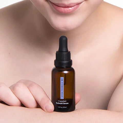 A person with bare shoulders holds an Open Formula Retinoid Serum bottle against their arm, highlighting its skin renewal benefits, with part of their face and a gentle smile visible.