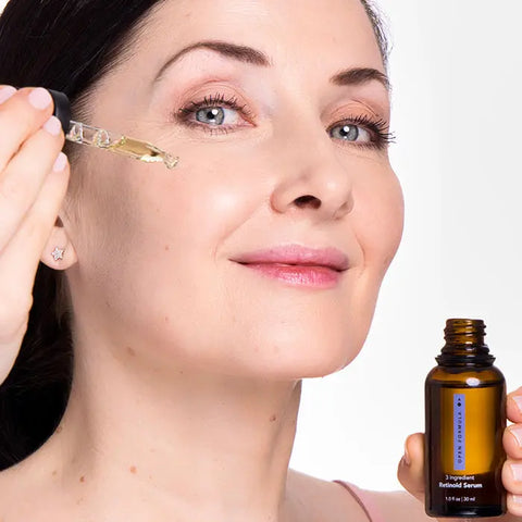 A woman with fair skin smiles slightly as she applies a drop of Open Formula Retinoid Serum to her cheek using a dropper. In the foreground, she holds the brown bottle with a blue label against a plain white background.