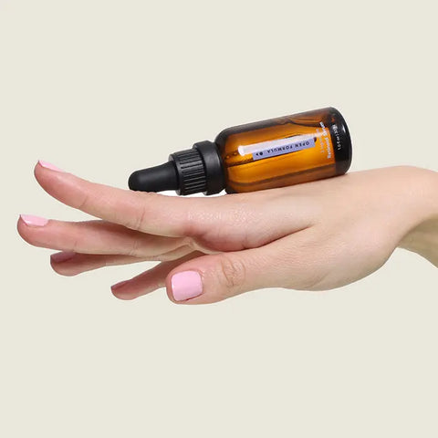 A hand with light pink nail polish balances a brown glass dropper bottle of Open Formula Retinoid Serum on the back of the fingers against a beige background, emphasizing this skin-renewing formula.