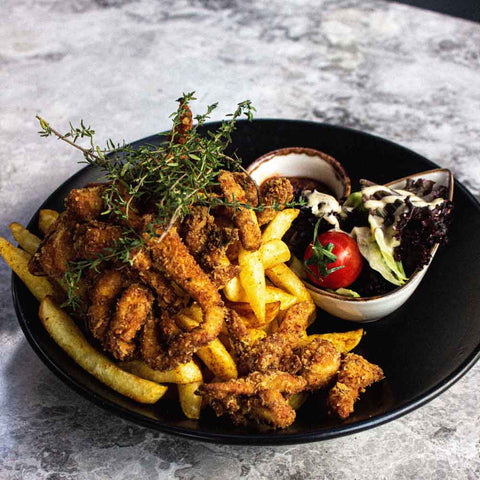A black plate features crispy fried chicken strips seasoned with Gustus Vitaes Air Fryer Kitchen Hero Seasoning Set, atop golden fries, garnished with fresh herbs, plus a lettuce and cherry tomato side salad with creamy dressing.
