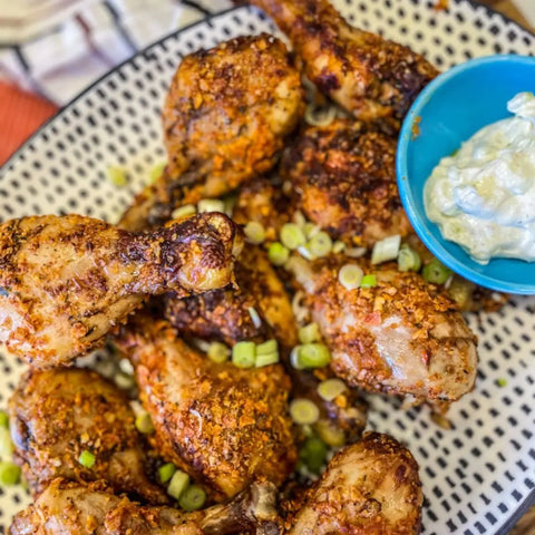 A plate of baked chicken drumsticks, garnished with green onions, seasoned using Gustus Vitae’s Air Fryer Kitchen Hero Seasoning Set - Artisanal Spice Blends Six Pack, served with creamy dipping sauce on the side.