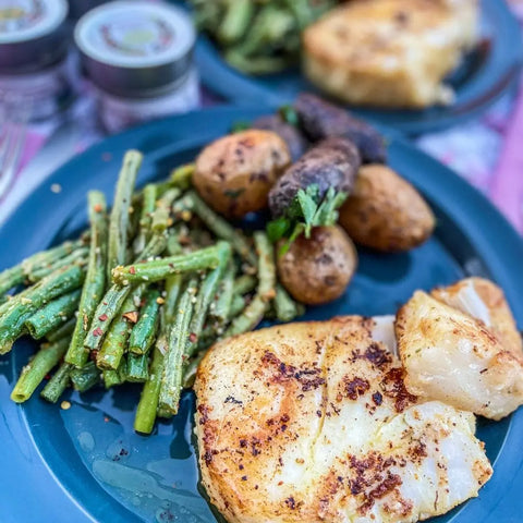 A blue plate features grilled white fish, green beans seasoned with Gustus Vitae’s Air Fryer Ultimate Veggies Seasoning Set, roasted baby potatoes, and skewered veggies. Another similar plate is blurred in the background.