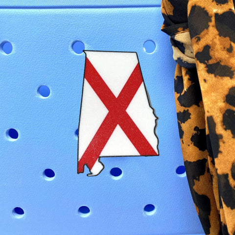 A Boglets USA Flag Charm by Chatelet Manufacturing, shaped like Alabama with a red X flag design, sits on a light blue perforated surface, while leopard print fabric hangs to the right. Compatible with Bogg Bags and more.