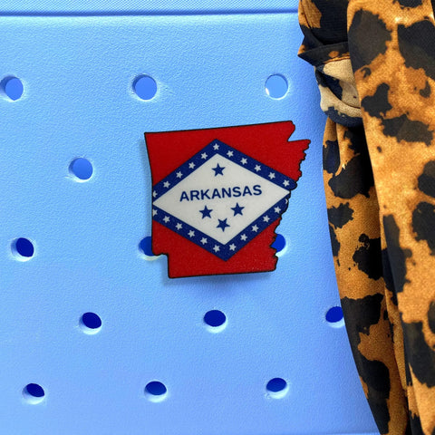 A Chatelet Manufacturing Boglets USA Flag Charm, designed for Bogg Bags and other totes, is displayed on a light blue surface near leopard print fabric and additional Bogg Bag charms.