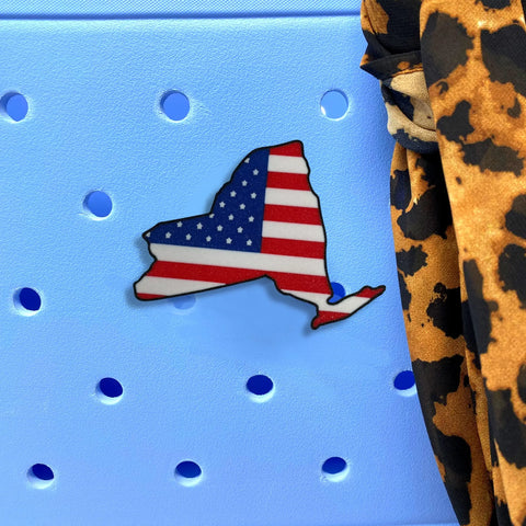 The Chatelet Manufacturing Boglets USA Flag Charm, shaped like New York State, is displayed on a light blue pegboard beside leopard print fabric, adding patriotic flair to Bogg Bags and other totes.