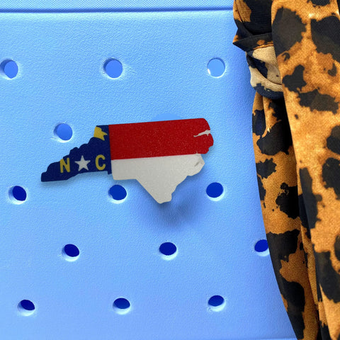 A Boglets USA Flag Charm by Chatelet Manufacturing is shown attached to a light blue perforated tote bag, with part of a leopard print fabric visible on the right. Perfect for displaying patriotism on Bogg Bags and other compatible totes.
