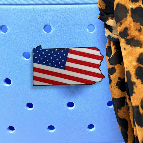 The Chatelet Manufacturing Boglets USA Flag Charm, a decorative pin for Bogg Bags and other totes, is shown attached to a light blue tote with round holes. Leopard print fabric is displayed nearby.