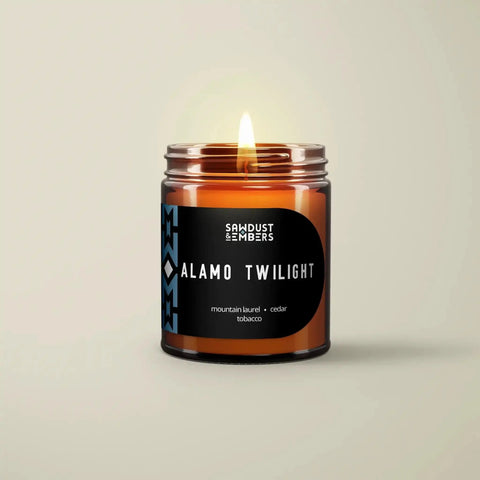 A lit Alamo Twilight - Signature Candle from Sawdust & Embers glows in an amber jar, featuring Texas-inspired scents of mountain laurel, cedar, and tobacco. Its label stands out with a black background and blue accents.