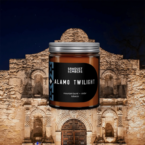 A brown Sawdust & Embers Alamo Twilight - Signature Candle sits before the illuminated Alamo Mission at night, capturing the Essence of Texas with mountain laurel, cedar, and tobacco notes.