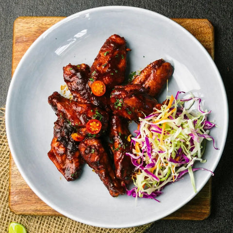 A white plate with barbecue chicken, garnished with cherry tomatoes, herbs, and artisan spices from the Gustus Vitae All American Wings Seasoning Set, is served with colorful shredded cabbage and carrots on a wooden board.