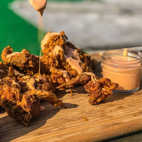 A close-up of crispy wings on a wooden board, drizzled with creamy sauce and served with Gustus Vitae’s All American Wings Seasoning Set for dipping. The blurred background features green and gray tones.