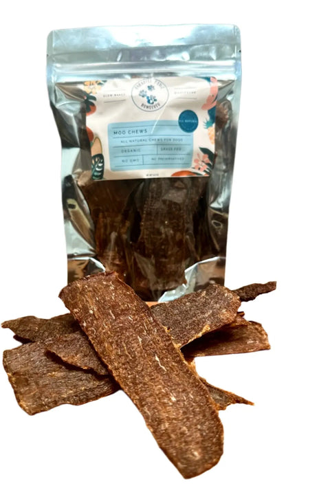 A silver resealable Moo Chews bag from Dogtown Bakery stands behind pieces of dried beef jerky made from grass-fed beef. The packaging features blue and white branding with floral accents.