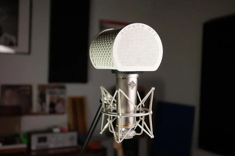 The Alpaca Mic, a large silver condenser microphone with a white pop filter, is set up on a stand in a recording room. The out-of-focus background shows shelves, equipment, and acoustic panels for professional-quality sound.
