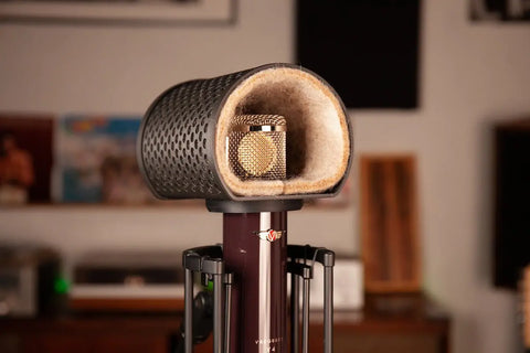 The Alpaca Mic, a gold-colored condenser microphone by Alpaca Mic, sits in a black foam-lined noise isolation shield on a stand in a recording studio with blurred shelves and equipment, providing professional sound quality.