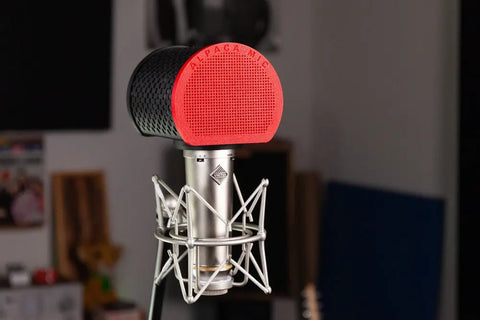 A close-up of the Alpaca Mic condenser microphone with a red Alpaca Mic filter featuring alpaca felt lining in a shock mount, set in a recording studio with blurred equipment and wall panels in the background for professional sound.