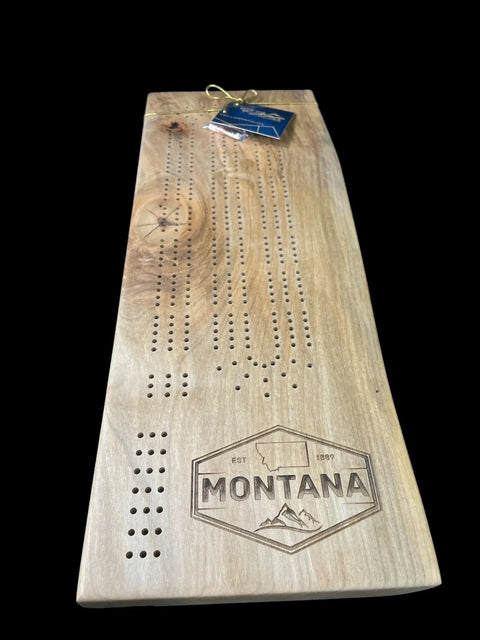 The B. James Design & Woodwork Ambrosia Maple Cribbage Board features a natural finish, Montana Emblem engraving with mountain design, peg holes, and a blue gift tag on top—all shown against a solid black background.