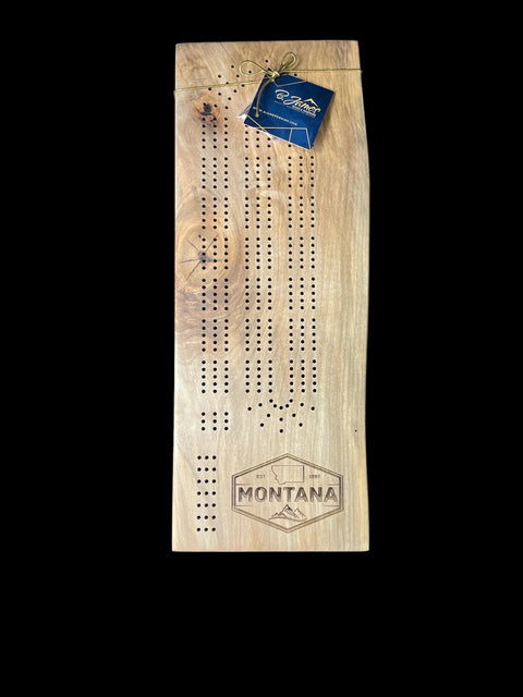 The Ambrosia Maple Cribbage Board With Montana Emblem Engraving by B. James Design & Woodwork features peg holes, a compass engraving, and mountain design on a black background, topped with a blue gift tag.