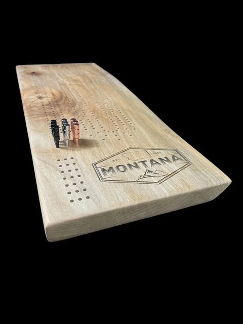 The Ambrosia Maple Cribbage Board With Montana Emblem Engraving by B. James Design & Woodwork features metal pegs and mountain artwork on a black backdrop—an ideal piece for Montana lovers.