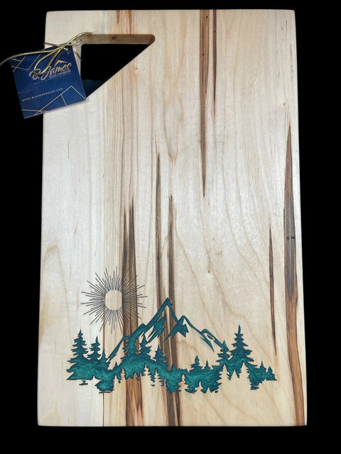 The Emerald Epoxy Inlay Ambrosia Maple Serving Tray by B. James Design & Woodwork features a triangular handle cutout and an engraved design of green trees, mountains, and a sun in the bottom left corner, with a branded tag on the handle.
