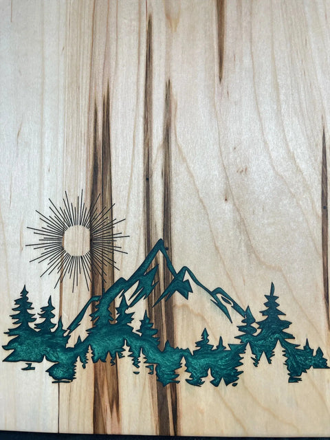 Handcrafted by B. James Design & Woodwork, the Emerald Epoxy Inlay Ambrosia Maple Serving Tray features a natural wooden grain with a sun, mountains, and green-accented pine trees, complemented by a unique emerald epoxy inlay for added charm.