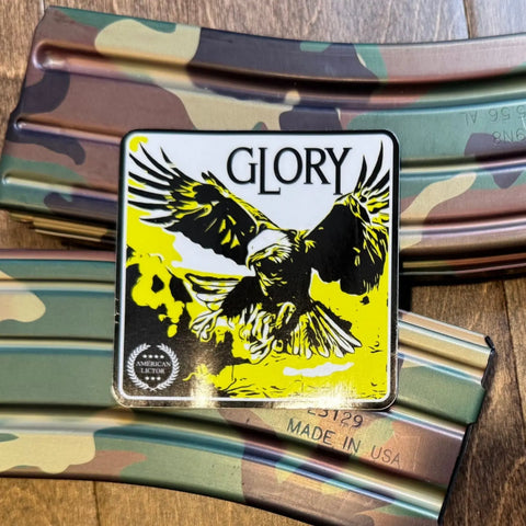 A rectangular Sticker: Bald Eagle - Glory from American Lictor, with an eagle and GLORY, rests on camouflage-patterned rifle magazines. Its bold black and yellow design is ideal for patriotic or country-themed decor.
