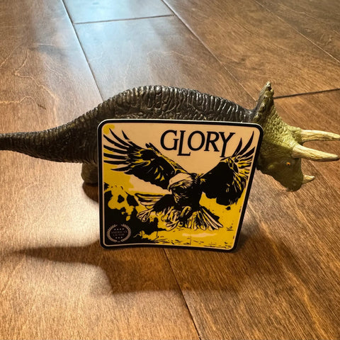 A toy triceratops stands on a wooden floor, with an American Lictor Sticker: Bald Eagle - Glory featuring a black and yellow eagle with outstretched wings on its side, adding patriotic flair to the playful scene.