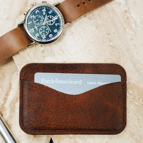 The Rogue Industries Bison Card Case, handcrafted in Maine from brown American Bison leather, holds a BankAmericard and sits on a light marble surface beside a matching brown leather-strap wristwatch.