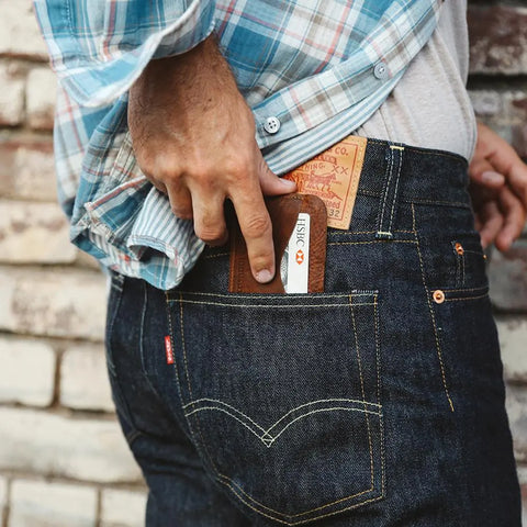 Wearing a plaid shirt and jeans, someone slips a Rogue Industries Bison Card Case—crafted from brown American Bison leather and made in Maine—holding a FISKC-labeled card into their back pocket, with a brick wall behind them.