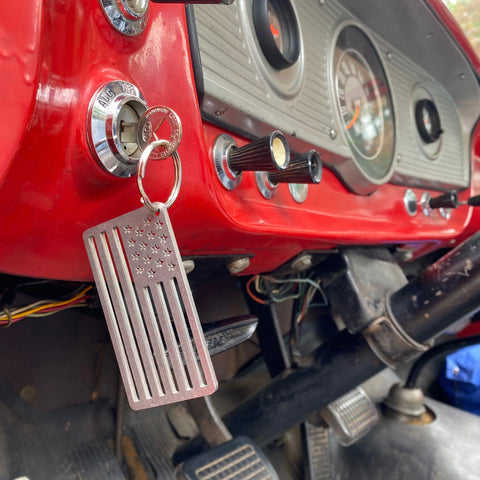 An American Flag Stainless Steel Key Chain by Arotags hangs from an ignition switch on a red vintage dashboard, adding a unique touch with its USA-made design and visible pedals and wires below.