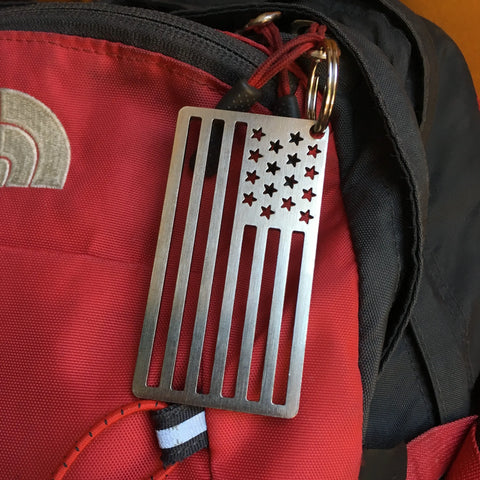 The Arotags American Flag Stainless Steel Key Chain, made in the USA with cutout stars and stripes, adds a unique vintage look when attached to the zipper of a red and black backpack.
