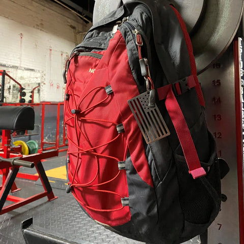 A red and black vintage-style backpack with an Arotags American Flag Stainless Steel Key Chain, made in the USA, hangs on gym equipment among benches, weights, and red metal frames in a weightlifting area.