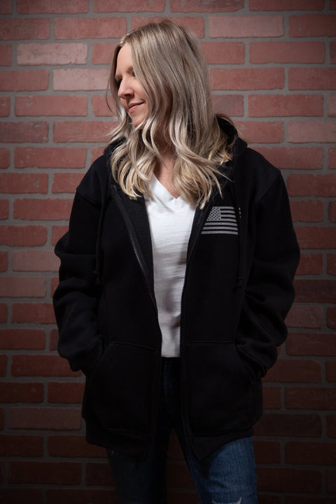 A blonde woman stands against a red brick wall, smiling to the side with her hands in the pockets of a Proudly USA American Flag - Zipper Hoodie Sweatshirt (Black), made in the USA and worn over a white shirt.