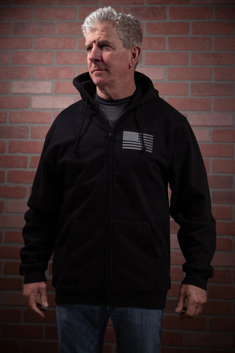 A gray-haired man wears the Proudly USA American Flag - Zipper Hoodie Sweatshirt (Black) while standing before a red brick wall, looking slightly to the side with a neutral expression.