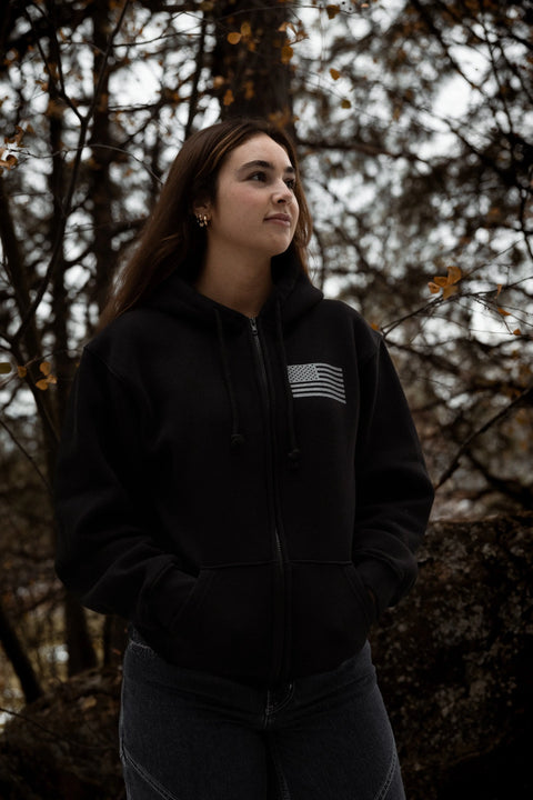 A young woman stands outdoors before trees, wearing the Proudly USA American Flag Zipper Hoodie Sweatshirt (Black) and dark jeans. With long brown hair and hands in her pockets, she looks off to the side with a slight smile.